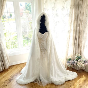 May include: A white bridal cape with a faux fur trim. The cape has a hood and is made of a soft, flowing fabric.