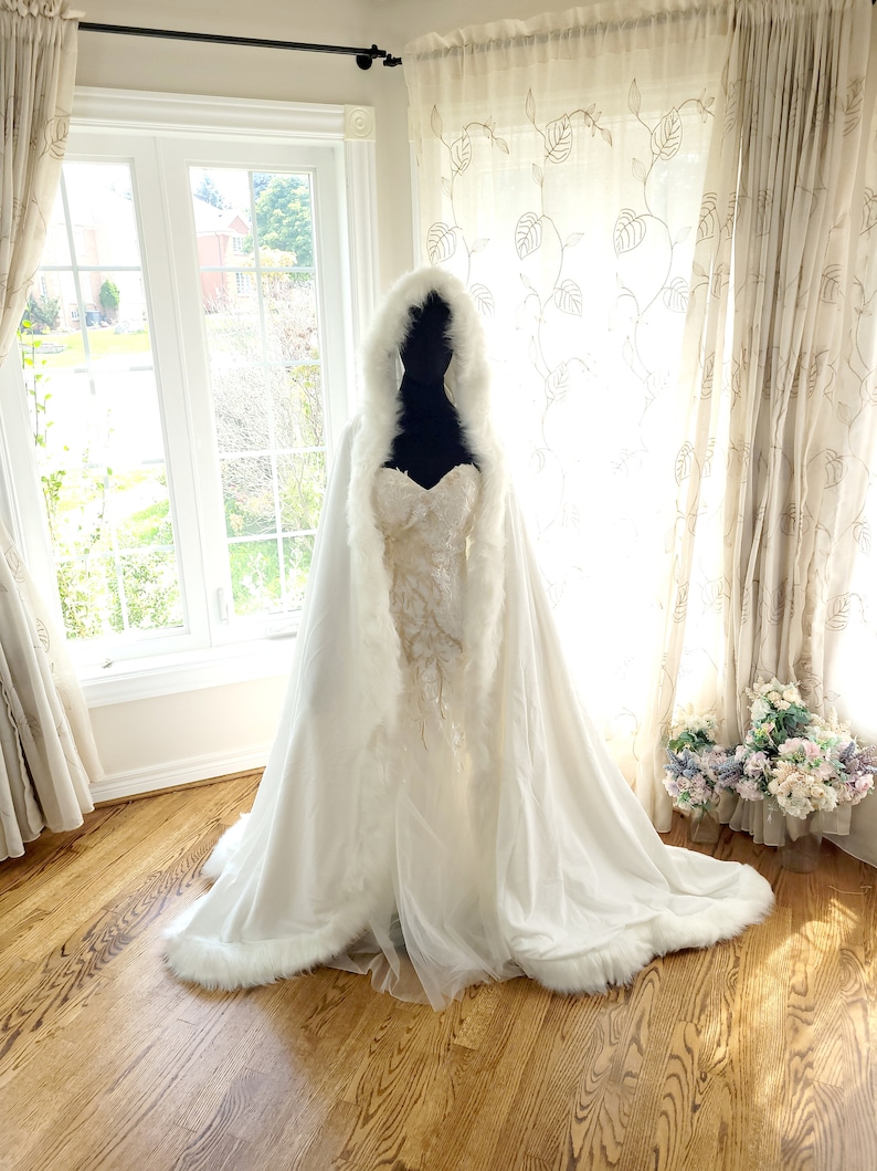 May include: A white bridal cape with a faux fur trim. The cape has a hood and is made of a soft, flowing fabric.