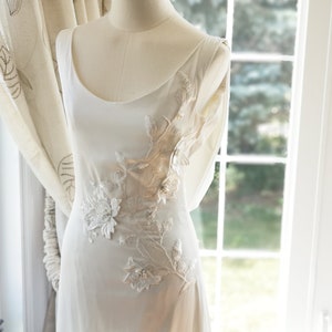 May include: A white sleeveless dress with a sheer overlay featuring delicate floral embroidery in shades of beige and white.