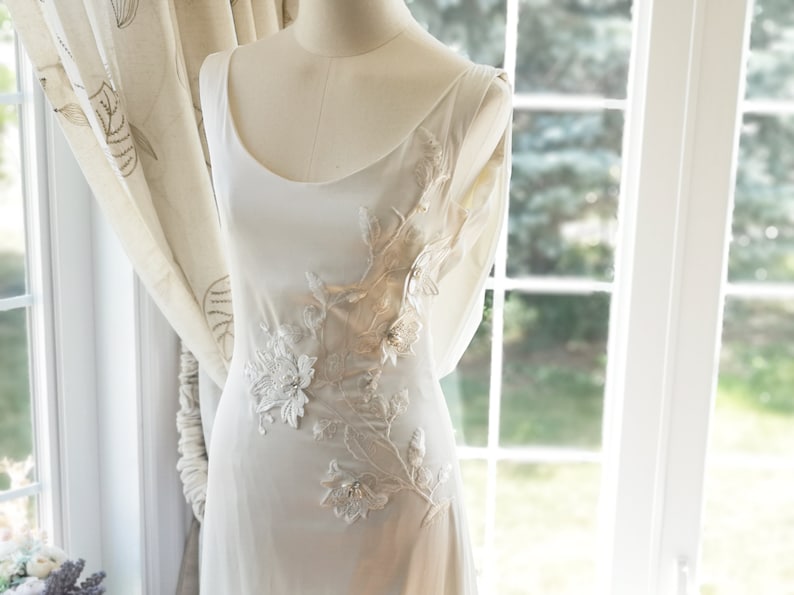 May include: A white sleeveless dress with a sheer overlay featuring delicate floral embroidery in shades of beige and white.