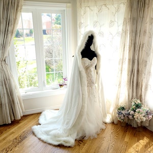 May include: A white bridal cape with a faux fur trim, worn over a white wedding dress with lace detailing. The cape has a hood and is draped over a mannequin in a room with curtains and a window.