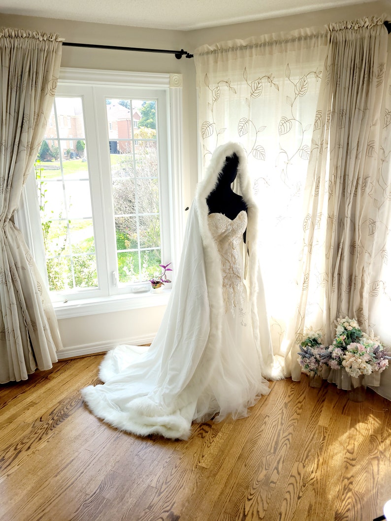 May include: A white bridal cape with a faux fur trim, worn over a white wedding dress with lace detailing. The cape has a hood and is draped over a mannequin in a room with curtains and a window.