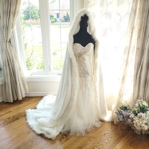 May include: A white bridal cape with a faux fur trim. The cape has a hood and is worn over a white wedding dress.