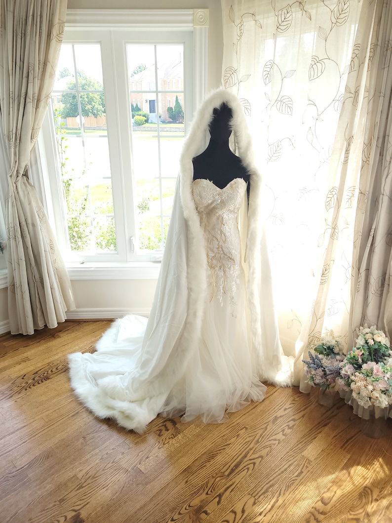 May include: A white bridal cape with a faux fur trim. The cape has a hood and is worn over a white wedding dress.