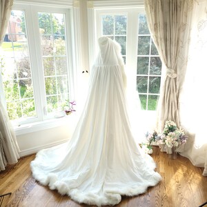 May include: A white hooded cape with a faux fur trim. The cape is laying on a wooden floor in front of a window.