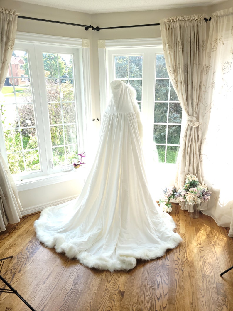May include: A white hooded cape with a faux fur trim. The cape is laying on a wooden floor in front of a window.