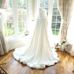 May include: A white hooded cape with a faux fur trim. The cape is draped over a mannequin and is displayed in a room with large windows and white curtains.