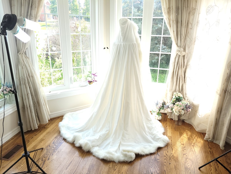 May include: A white hooded cape with a faux fur trim. The cape is draped over a mannequin and is displayed in a room with large windows and white curtains.