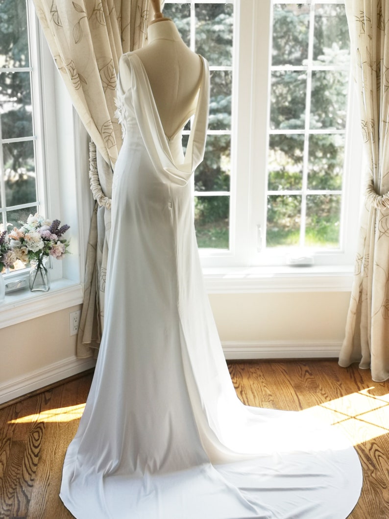 May include: A white wedding dress with a low back and a flowing train. The dress is made of a smooth, silky fabric and has a simple, elegant design.
