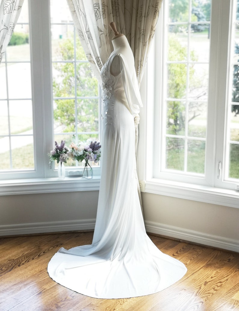 May include: A white wedding dress with a fitted bodice and a flowing skirt. The dress has a lace overlay on the bodice and a small train. The dress is displayed on a mannequin against a white background.