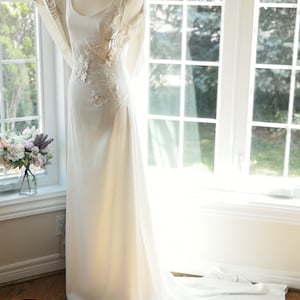 May include: A white wedding dress with a scoop neckline and a fitted bodice. The dress has a flowing skirt and a train. The bodice is adorned with delicate floral embroidery. The dress is displayed on a mannequin against a window background.