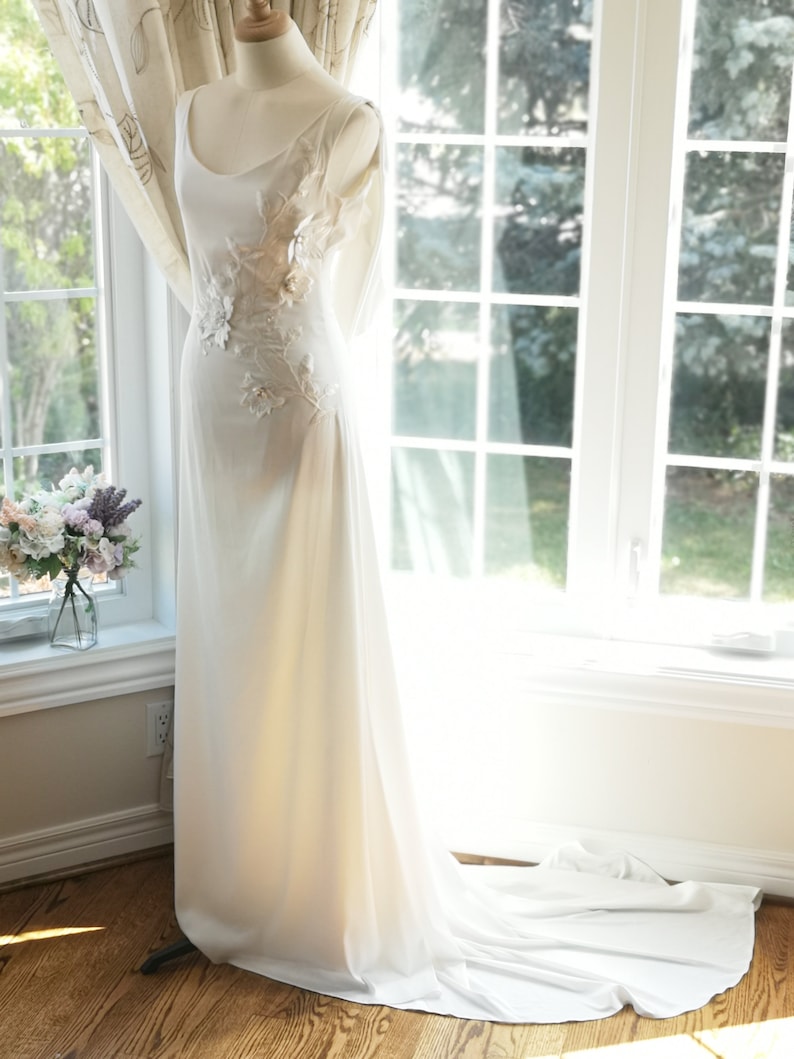 May include: A white wedding dress with a scoop neckline and a fitted bodice. The dress has a flowing skirt and a train. The bodice is adorned with delicate floral embroidery. The dress is displayed on a mannequin against a window background.