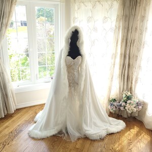 May include: A white faux fur bridal cape with a hood, trimmed with white fur. The cape is draped over a white wedding dress with lace detailing.