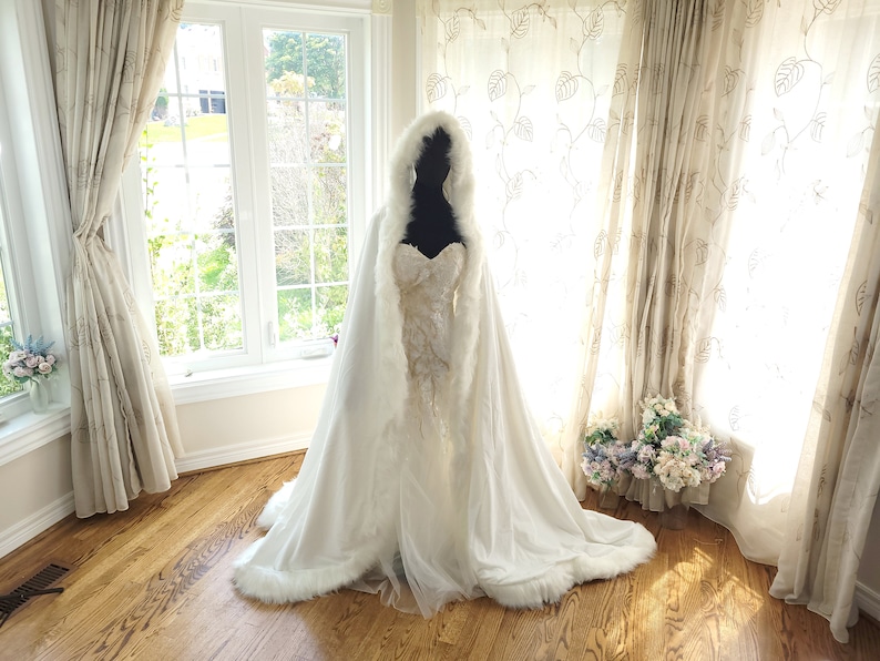 May include: A white faux fur bridal cape with a hood, trimmed with white fur. The cape is draped over a white wedding dress with lace detailing.