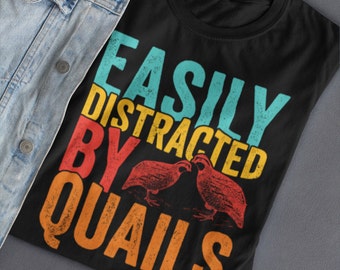 May include: Black t-shirt with the text "EASILY DISTRACTED BY QUAILS" in teal, yellow, red, and orange. Two quail illustrations are below the text. The shirt is on a gray surface with a denim jacket.