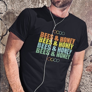 Peut inclure: T-shirt noir avec l'inscription "BEES & HONEY" dans un style rétro. Le texte est superposé et de couleurs orange, jaune, sarcelle et vert. Le t-shirt comporte également un motif en nid d'abeille.