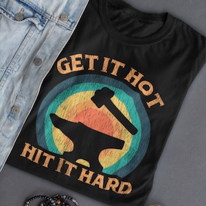 May include: Black t-shirt with a vintage-style graphic of a hammer and anvil. The graphic has a rainbow color scheme and the text "Get it Hot Hit it Hard".