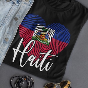 Op de afbeelding: Zwart T-shirt met een wit hartvormig vingerafdrukdesign met de kleuren van de Ha&iuml;tiaanse vlag: blauw, rood en groen. Het woord "Ha&iuml;ti" staat in wit onder het hart.