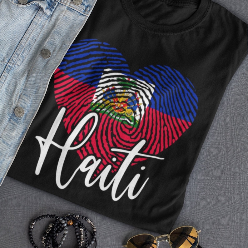 Op de afbeelding: Zwart T-shirt met een wit hartvormig vingerafdrukdesign met de kleuren van de Ha&iuml;tiaanse vlag: blauw, rood en groen. Het woord "Ha&iuml;ti" staat in wit onder het hart.