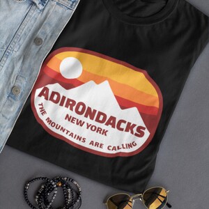 May include: Black t-shirt with a red, orange, and yellow graphic of mountains and the text "Adirondacks The Mountains Are Calling New York".