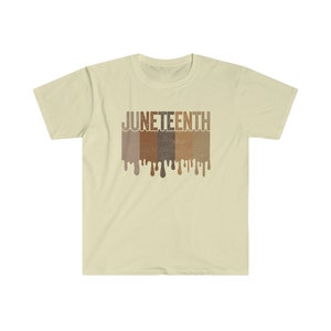 May include: A beige t-shirt with a graphic print of the word "JUNETEENTH" in brown, tan, and dark brown letters. The letters are dripping with paint, creating a dripping effect.