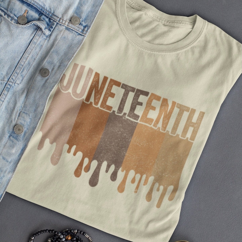 May include: A light beige t-shirt with the word "Juneteenth" printed in brown, orange, and beige letters. The letters are dripping with paint, creating a colorful and abstract design.