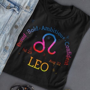 Op de afbeelding: Zwart T-shirt met een regenboogkleurig Leeuw-sterrenbeeld en de tekst "Proud. Bold. Ambitious. Confident" rond het teken. De data "Jul 23" en "Aug 22" zijn ook inbegrepen.