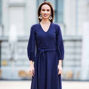 May include A woman wearing a navy blue midi dress with a self-tie belt and long sleeves. She is wearing burgundy ankle boots.
