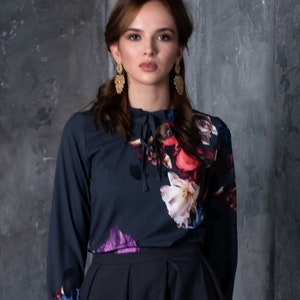 May include: A woman wearing a dark blue blouse with a floral print and a dark blue skirt. The blouse has a tie at the neck and long sleeves.
