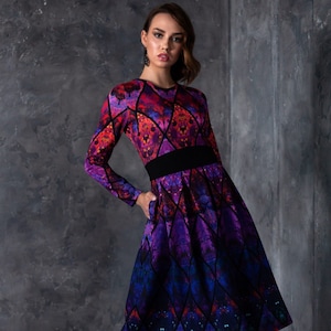 Può includere: Una donna indossa un abito a maniche lunghe con un motivo geometrico colorato. L'abito ha una cintura nera e una gonna svasata. Indossa collant neri e stivali con tacco bordeaux.