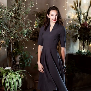 May include A woman wearing a dark gray knee-length dress with a v-neck and a flared skirt. She is wearing black tights and gray ankle boots. The woman is standing in a room with plants and string lights.