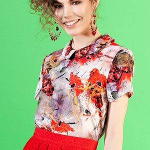 May include: A woman with curly brown hair wearing a white blouse with a floral print and a red skirt. She is wearing red earrings.