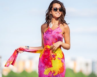 May include: A woman wearing a pink, yellow, and purple maxi dress with a tie at the waist. She is wearing sunglasses and has long brown hair.
