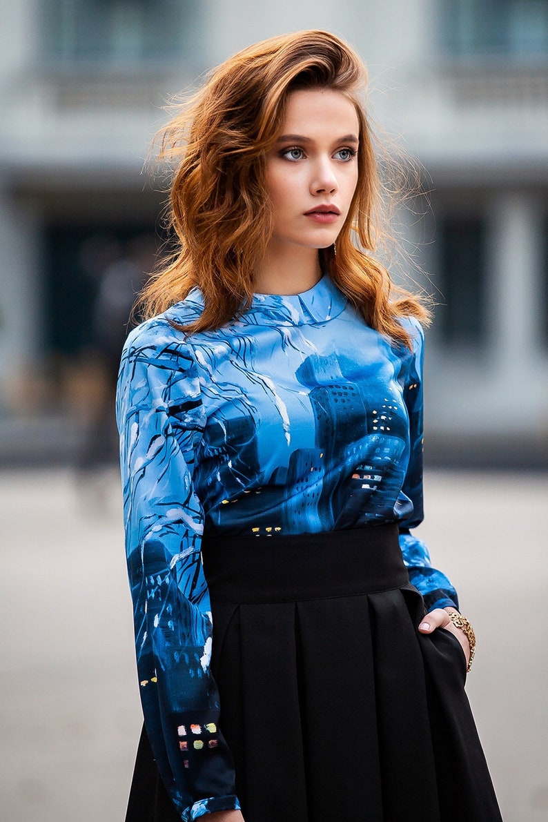 K&ouml;nnte beinhalten: Eine Frau tr&auml;gt eine blau-schwarze gemusterte Bluse mit einem schwarzen Rock. Die Bluse hat einen Stadtbild-Druck.