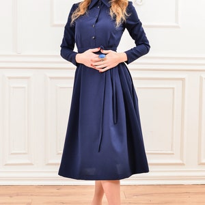 Pu&ograve; includere: Un abito midi blu navy con maniche lunghe, chiusura con bottoni sul davanti e una cintura annodata in vita. L'abito ha una gonna leggermente svasata ed &egrave; indossato con tacchi neri.