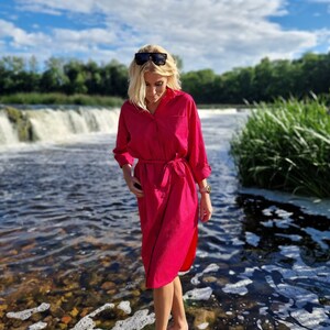 May include: A woman wearing a long-sleeved, fuchsia-colored dress with a belt, standing in a river. The dress has a button-down design. Sunglasses rest on her head. The background features a waterfall, trees, and a blue sky with clouds.