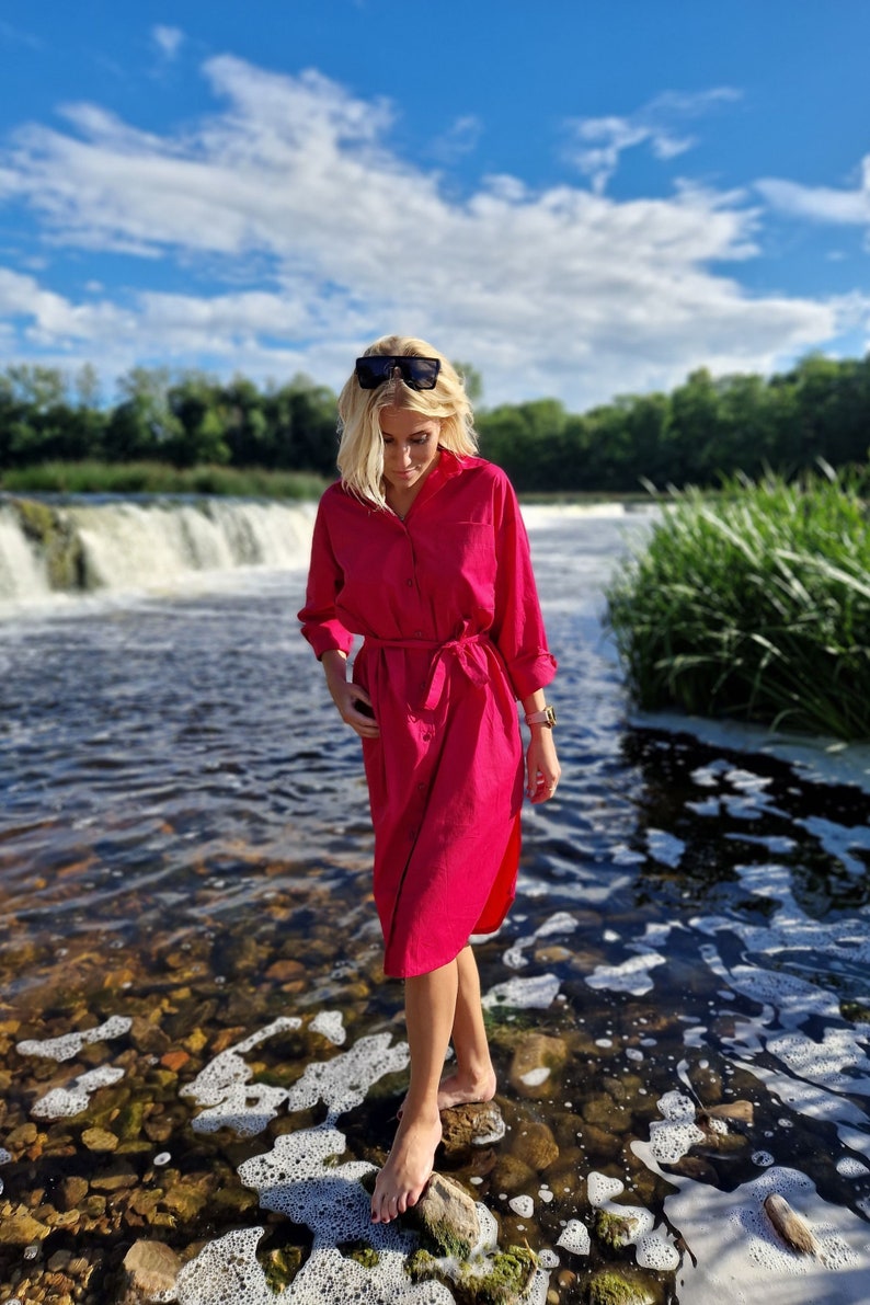 May include: A woman wearing a long-sleeved, fuchsia-colored dress with a belt, standing in a river. The dress has a button-down design. Sunglasses rest on her head. The background features a waterfall, trees, and a blue sky with clouds.