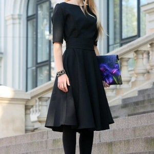 Op de afbeelding: Een vrouw in een zwarte jurk met een getailleerd bovenstuk en een volle rok. Ze draagt een bloemenclutch en zwarte panty's en hakken.