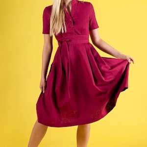May include A woman wearing a burgundy midi dress with a tie belt and short sleeves. She is standing on a yellow background and wearing blue heels.
