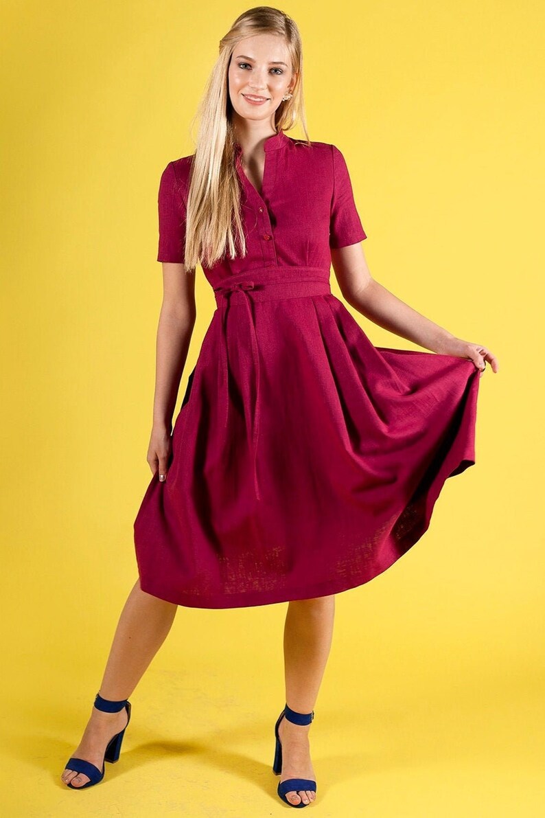 May include A woman wearing a burgundy midi dress with a tie belt and short sleeves. She is standing on a yellow background and wearing blue heels.