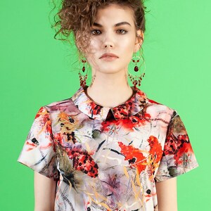May include: A woman with curly brown hair wearing a white collared blouse with a red, orange, and yellow floral print. She is also wearing a red skirt and red dangle earrings.