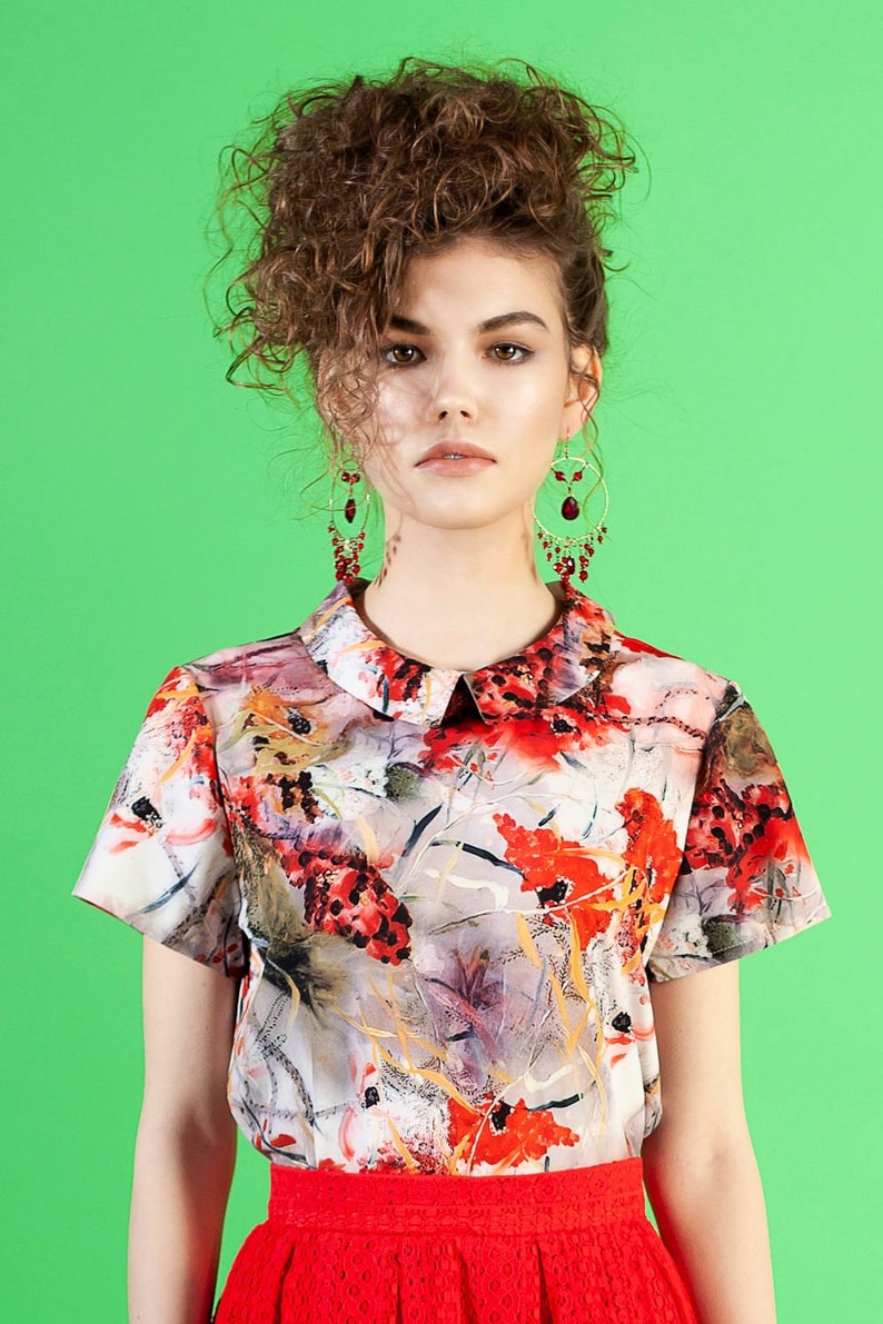 May include: A woman with curly brown hair wearing a white collared blouse with a red, orange, and yellow floral print. She is also wearing a red skirt and red dangle earrings.