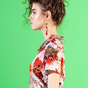 May include: A woman with curly brown hair wearing a white and red floral print blouse with a red skirt and red earrings.