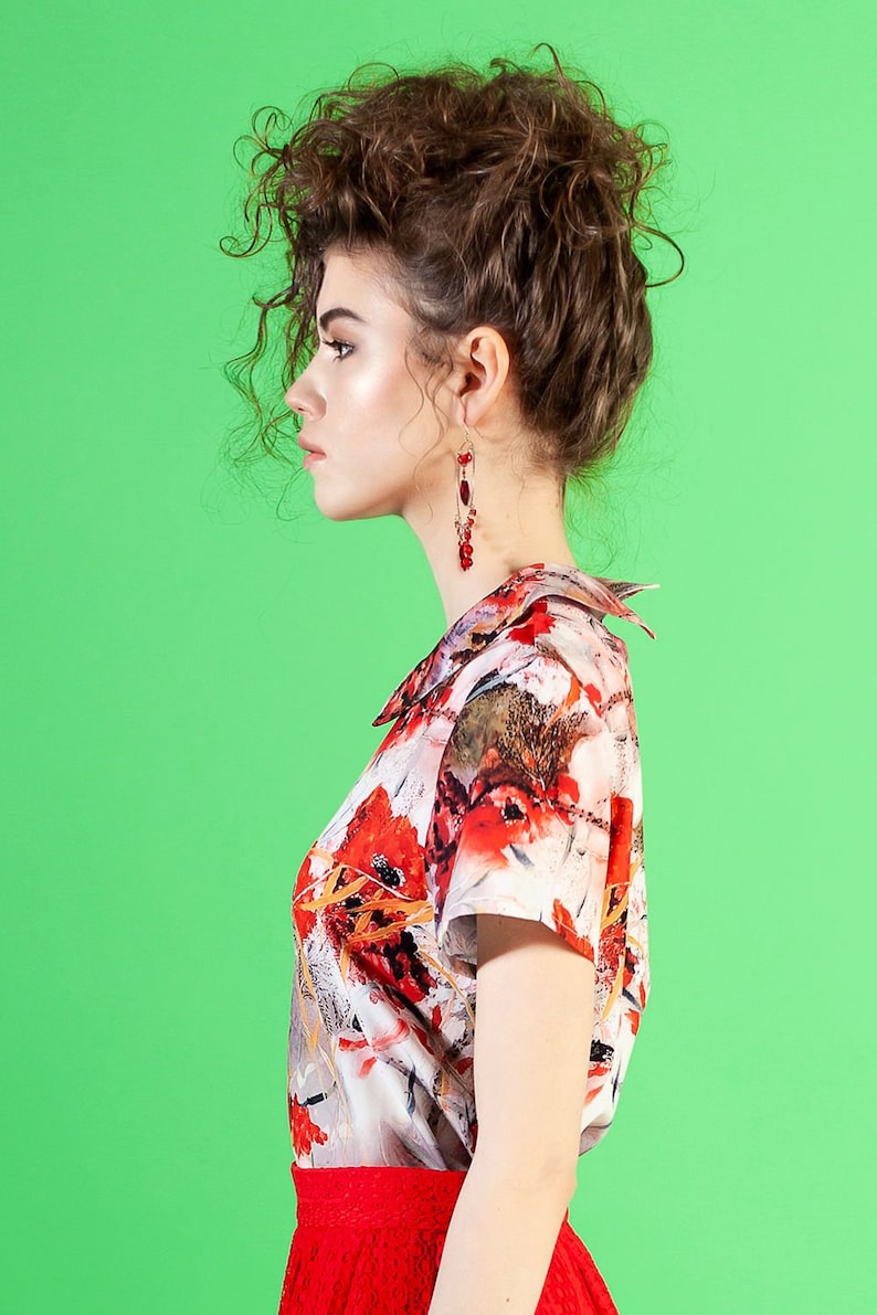 May include: A woman with curly brown hair wearing a white and red floral print blouse with a red skirt and red earrings.