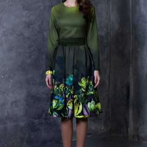 May include: A woman wearing a green dress with a floral print. The dress has a fitted bodice and a full skirt. The woman is standing in front of a gray textured wall.