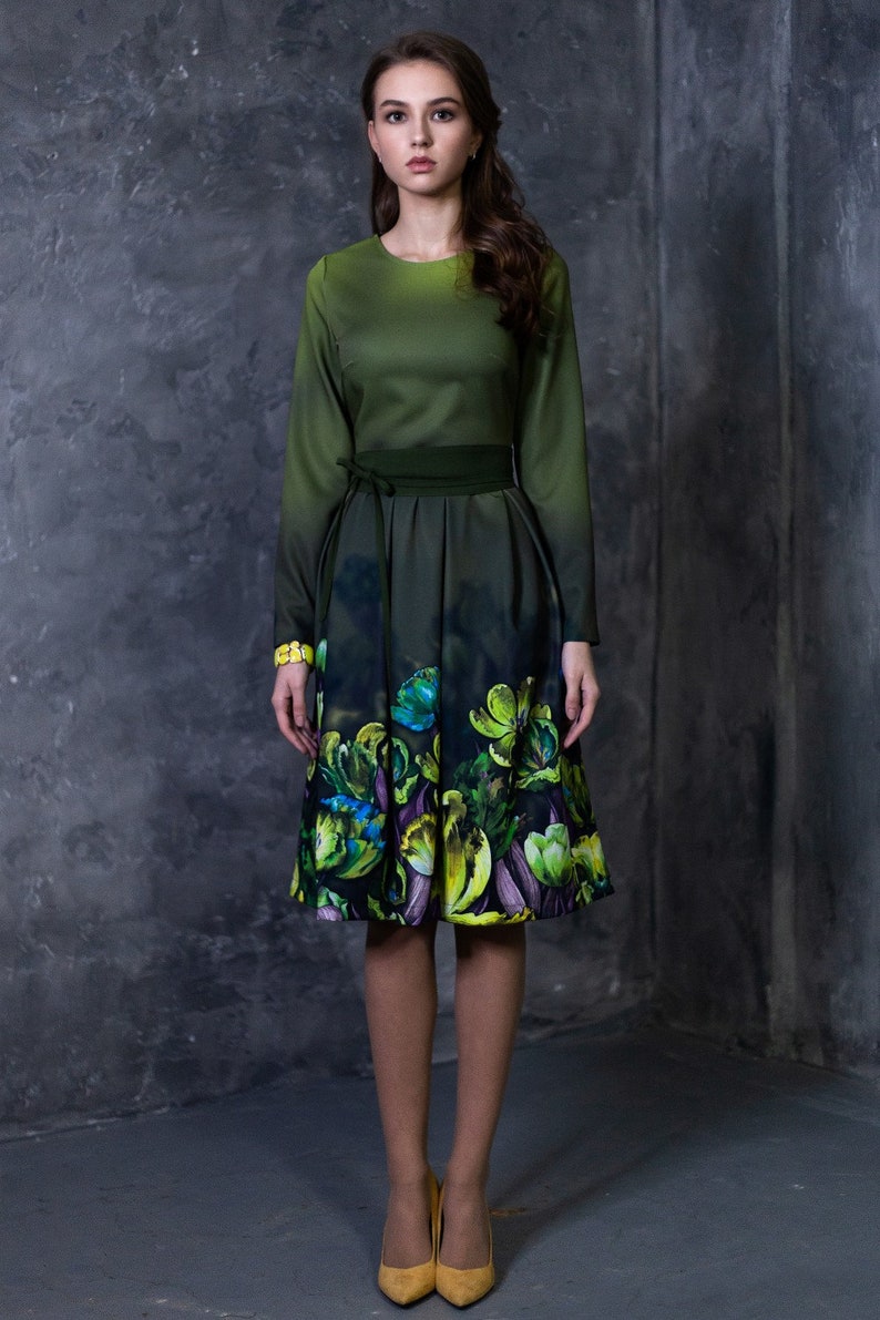 May include: A woman wearing a green dress with a floral print. The dress has a fitted bodice and a full skirt. The woman is standing in front of a gray textured wall.