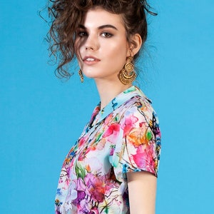 May include: A woman with curly brown hair wearing a blue skirt and a white blouse with a colorful floral print. She is wearing gold earrings.
