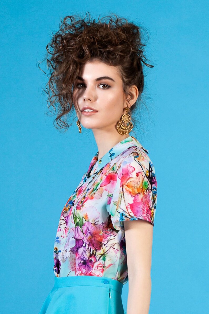 May include: A woman with curly brown hair wearing a blue skirt and a white blouse with a colorful floral print. She is wearing gold earrings.