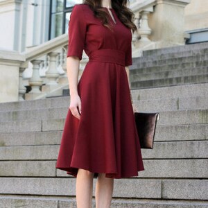 Pode incluir: Uma mulher vestindo um vestido vinho com um corpete justo e uma saia rodada. Ela est&aacute; em p&eacute; em uma escada e segurando uma embreagem marrom.
