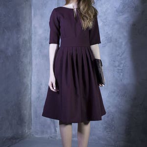 May include: A woman wearing a burgundy dress with a pleated skirt and a small clutch. She is standing in front of a gray wall.